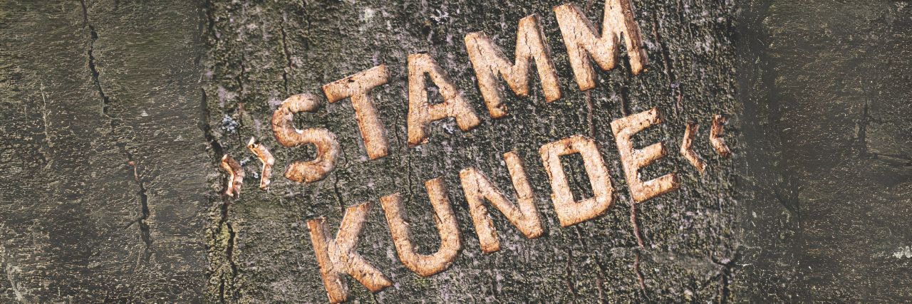 In der Rinde eines dunklen Baumstamms ist das Wort Stammkunde eingeritzt