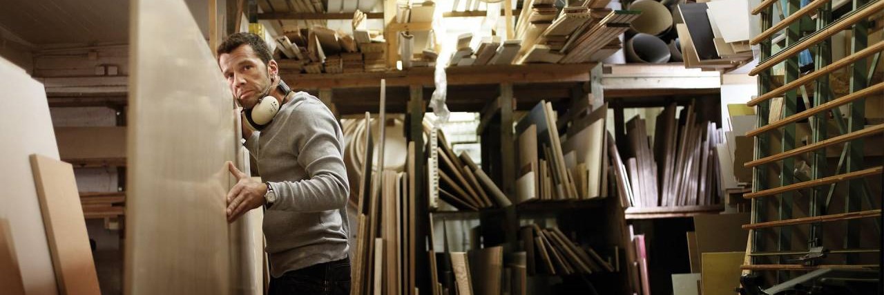 Ein Tischler in einer Holzwerkstatt fässt eine senkrecht stehende Holzplatte an und schaut über den Betrachter hinweg in die Ferne